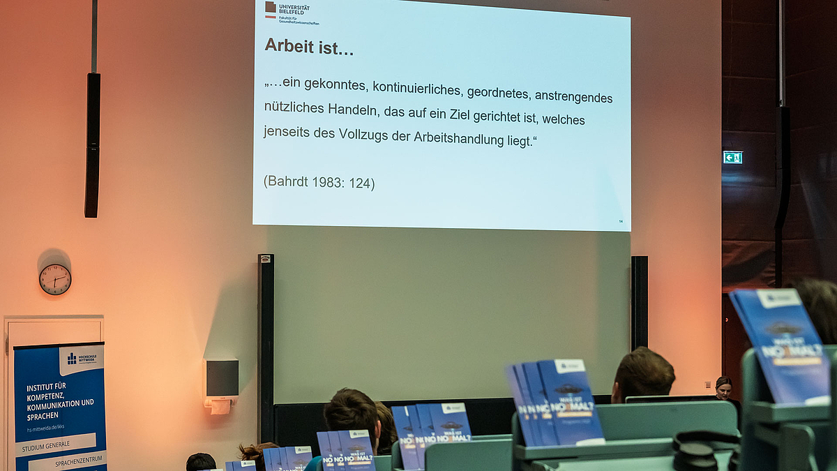 Blick über die Köpfe von Zuhörenden im Hörsaal hinweg auf eine projizierte Folie mit dem Zitat von Hans Paul Bahrdt aus dem Jahr 1983: „Arbeit ist ein gekonntes, kontinuierliches, geordnetes, anstrengendes nützliches Handeln, das auch ein Ziel gerichtet ist, welches jenseits der Arbeitshandlung liegt.“