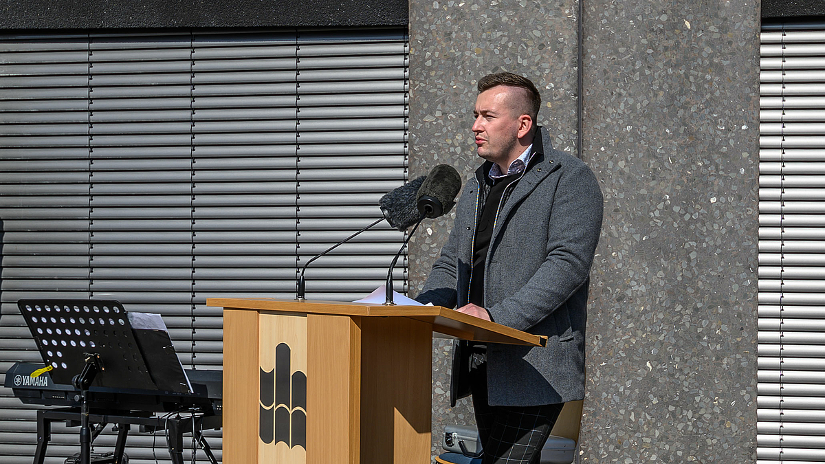 Studentisches Engagement in der Pandemie und für die Ukraine. Stura Geschäftsführer Gordon Guido Oswald: „Wir sind von einer Krise in die nächste gerutscht. Aber auch diese Hilfsbereitschaft macht unser Campusleben aus.“ Das Foto zeigt einen Mann am Rednerpult. Aus der Vordersite des Pults ist das Logo der Hochschule vollständig zu sehen.