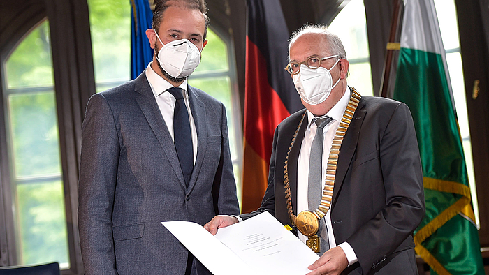 Das Foto zeigt zwei männliche Personen jeweils mit weißer FFP-Maske: links den Sächsischen Wissenschaftsminister Sebastian Gemkow, daneben den Mittweidaer Hochschulrektor Ludwig Hilmer. Dieser hält eine geöffnete Mappe mit der soeben unterzeichneten Vereinbarung zum Betrachtenden. Der Inhalt ist zu erkennen.