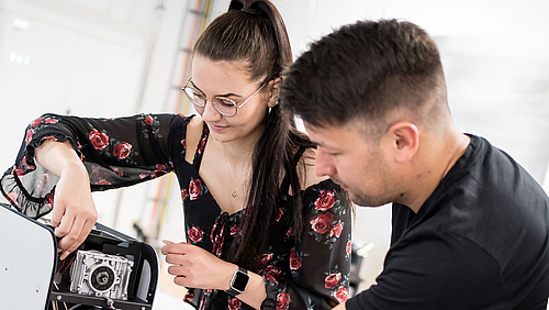 Eine Studentin arbeitet am Inneren eines technischen Geräts, der junge Dozent steht neben ihr zur Unterstützung bereit.