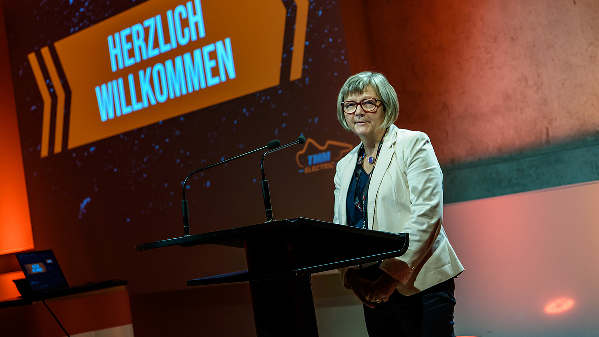 Auf dem Fotos ist eine Frau am Rednerpult zu sehen. Auf der Leinwand im Hintergrund ist "Herzlich willkommen" zu lesen.