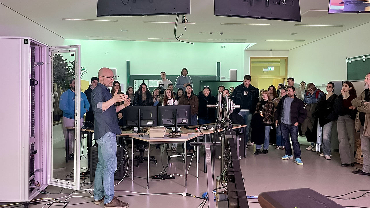 Ein Studioraum mit vielen stehenden jungen Menschen, die einem Dozenten zuhören.