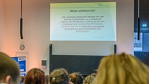 Blick von hinten über die Köpfe der Zuschauenden auf den Dozenten, der vor der Tafel steht. Über ihm zeigt die Projektion den Text: „Woran scheitern wir? Still, promoting collaboration between the wide diversity of stakeholders, as a fundamental aspect in the OH (One Health) approach, is still by far the most challenging factor in performing OH initiatives. Causes for that are the difficulties in promoting meaningful and equal participation from diverse actors.”