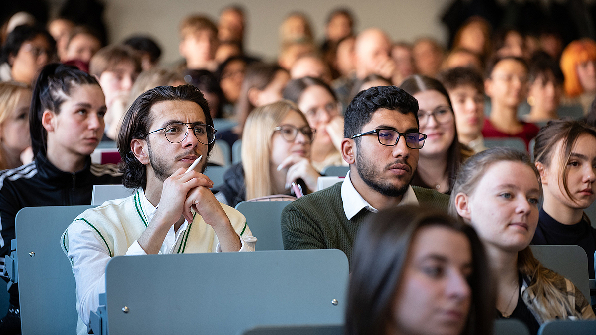 Zwei junge Männer sitzen auf einer Hörsaalbank und lauschen. Um sie herum sitzen weitere Studierende.