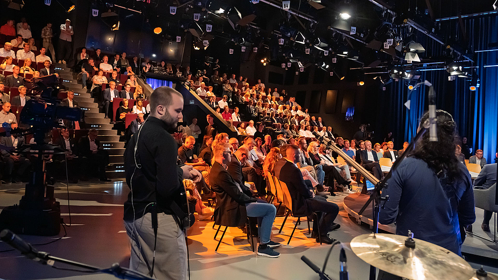 Volle Zuschauerränge im Fernsehstudio. Im Vordergrund sind von hinten zwei Musiker einer Band von hinten zu sehen.