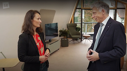 Die Journalistin aus dem Saarland und der Hochschulrektor aus Sachsen. Diana Kühner-Mert im Gespräch mit Professor Volker Tolkmitt (Screenshot „Im Wandel vereint“/SR/ARD-Mediathek) Eine weibliche und eine Person stehen sich im Gespräch in einem Raum mit einer großen Fensterfläche im Hintergrund gegenüber. in der oberen linken Bildecke ist das Sender-Logo "SR" zu sehen.