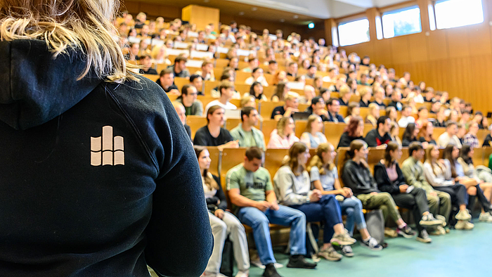 Die Bankreihen eines Hörsaals sind voll besetzt. Am linken Bildrand steht eine Person, die sich den Pesonen im Hörsaal zuwendet. Sie trägt einen blauen Kapuzenpullover mit dem Hochschullogo auf dem Rücken. 
