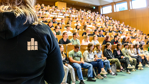 Die Bankreihen eines Hörsaals sind voll besetzt. Am linken Bildrand steht eine Person, die sich den Pesonen im Hörsaal zuwendet. Sie trägt einen blauen Kapuzenpullover mit dem Hochschullogo auf dem Rücken. 