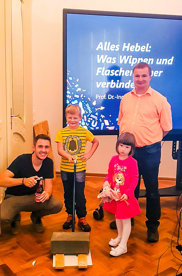 Das Foto zeigt zwei Kinder, Paul und Celina, zwischen zwei Erwachsenen vor einem angeschnittenen großen Monitor im Hintergrund: links knieend Robin Biebl, Kinderuni-Organisator, rechts Kinderuni-Dozent Prof. Uwe Mahn.