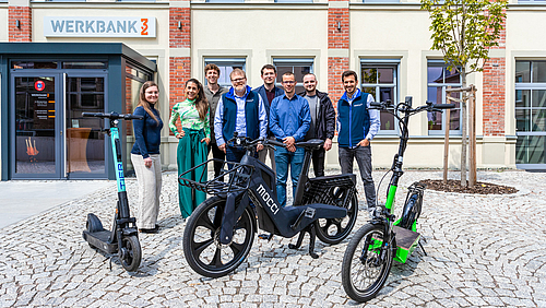 Das Foto zeigt eine Gruppe von 8 Personen auf einem gepflasterten Platz vor einem Gebäude stehend, das den Namen Werbank32 trägt. Vor der Gruppe stehen drei zweirädrige Fahrzeuge, zwischen zwei E-Scootern ein futuristisches Pedelec mit der Aufschrift mocci.