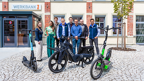 Das Team des Instituts für Energiemanagement der Hochschule Mittweida (ifem) und ihrer Kooperationspartner:innen mocci und blXmove stehen vor der Zukunft vernetzter Mobilität in der Hochschulstadt. (Foto: Erik Hilse) Das Foto zeigt eine Gruppe von 8 Personen auf einem gepflasterten Platz vor einem Gebäude stehend, das den Namen Werbank32 trägt. Vor der Gruppe stehen drei zweirädrige Fahrzeuge, zwischen zwei E-Scootern ein futuristisches Pedelec mit der Aufschrift mocci.