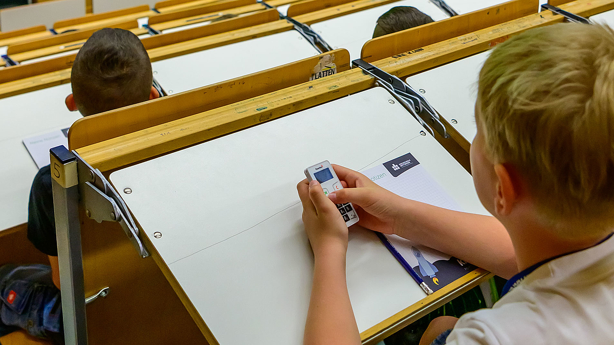 Das Foto zeigt ein Kind von hinten in einer Hörsaalbank, das ein ein Eingabegraät für eine drahtlose Abstimmung in der Hand hält.