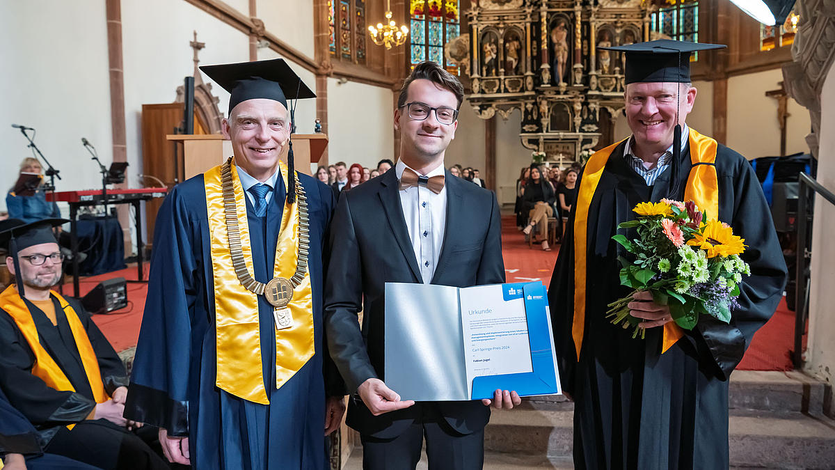 Jüngere männliche Person im Anzug mit Urkunde in der Hand steht zwichen zwei älteren männlichen Personen im Talar. Die rechte Person hat einen Blumenstrauß in der Hand.