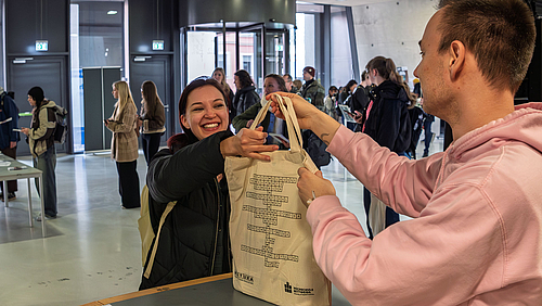 Eine strahlend lächelnde weibliche Person erhält aus den Händen einer männlichen Person einen Stoffbeute über einen Tresen gereicht. 
