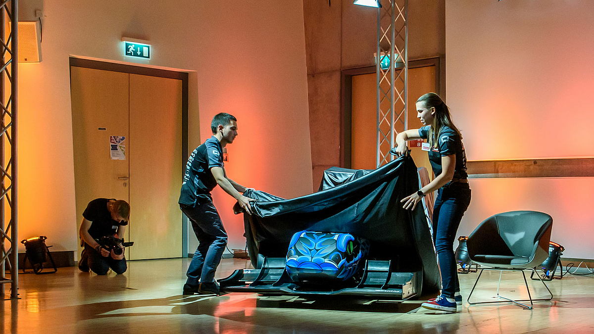 Das Foto zeigt den jungen Mann vom Aufmacherfoto und die junge Frau au der Talkrunde links und rechts vom Rennwagen stehend und die schwarze Plane vom Auto zu ziehen, um es zu enthüllen.