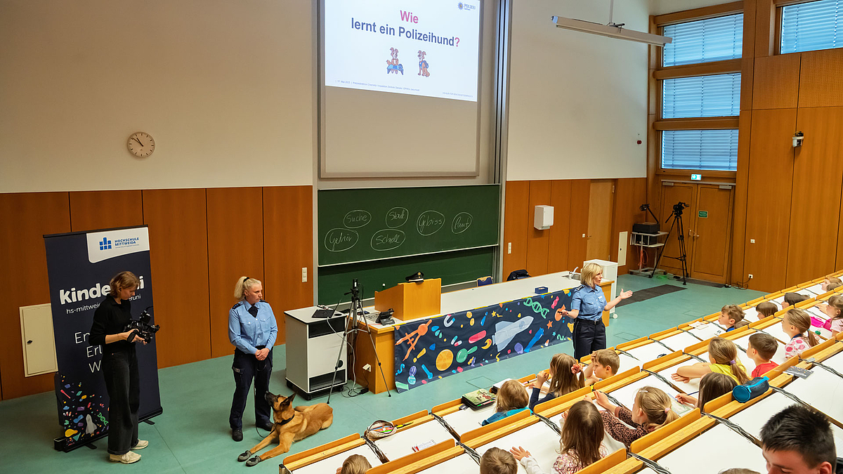 Blick über die Köpfe der Kinder hinweg auf Hundeführerin, Hund und Referentin. An der Wand im Hintergrund ist dei Folie projiiziert mit dem Text "Wie lernt ein Polizeihund?"