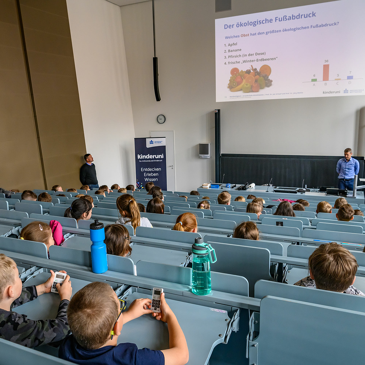 Blick über die Köpfe der Kinder hinweg auf die beiden Dozenten. An die Rückwand über ihnen ist eine Folie projiziert mit der Frage „Welches Obst hat den größten ökologischen Fußabdruck?“ und den Auswahlmöglichkeiten „Apfel, Banane, Pfirsich in der Dose, Frische Winter-Erdbeeren“.  Im Bildvordergrund sind zwei Kinder zu sehen, die jeweils ein mobiles Abstimmgerät in der Hand halten, das ähnlich aussieht wie ein Taschenrechner.