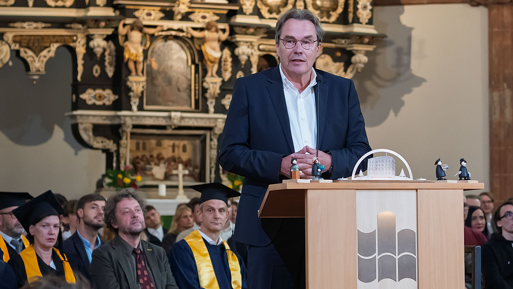 Männliche Person am Rednerpult, hiter ihr sitzen Professor:innen und Erstsemester im Altarraum