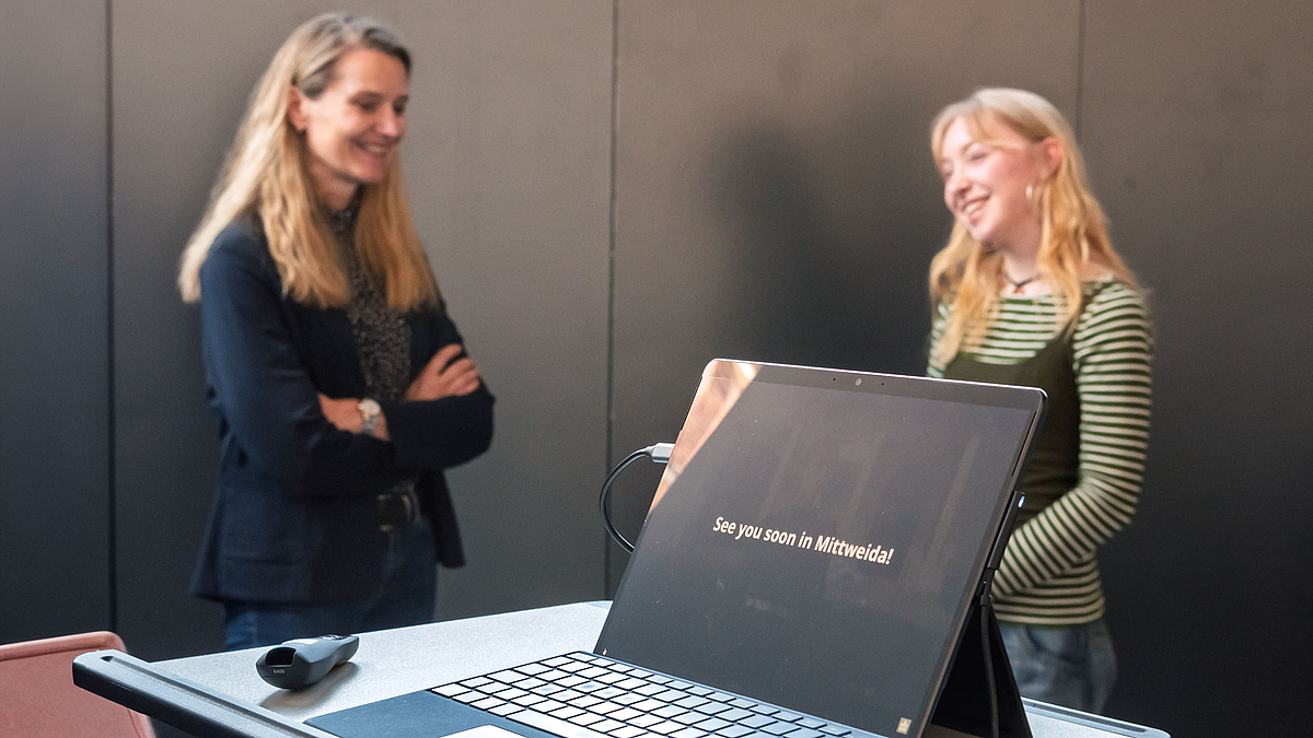 Ein aufgeklappter Laptop auf einem Pult. Auf dem Display ist der Satz: "See you soon in Mittweida" zu lesen. Im Hintergrund stehen zwei lächelnde weibliche Personen.