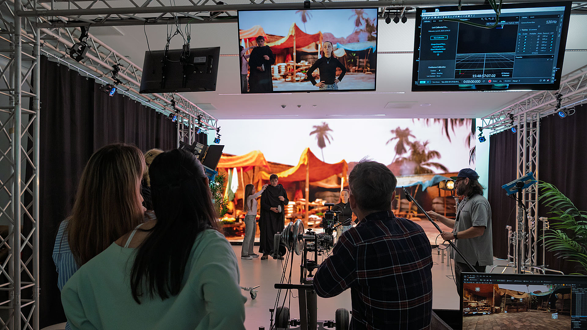 Studioraum mit Scheinwerfen und Monitoren. Personen im Bildvordergrund beobachten die vor einer raumhohen LED-Wand agierenden Personne. Die LED-Wand zeigt einen orientalischen Markt als Hintergrund für die Szene.