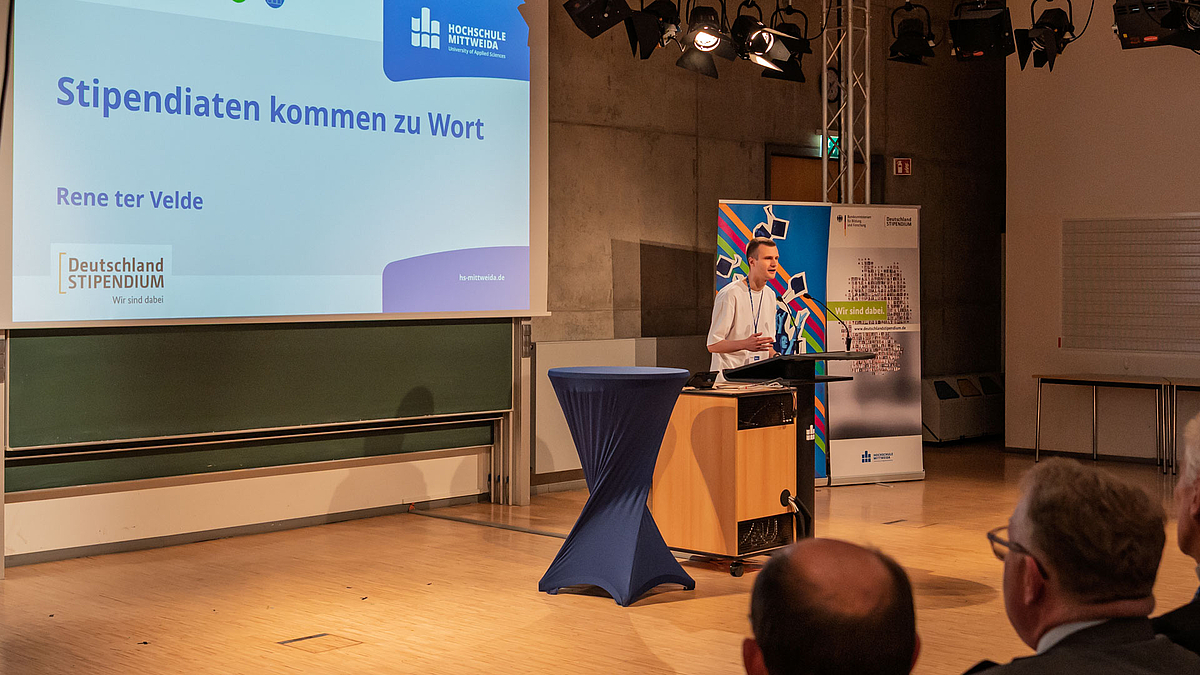Junger männlicher Redner am Pult. Auf der Leinwand ist zu lesen: "Stipendiaten kommen zur Wort. Rene der Velde"-