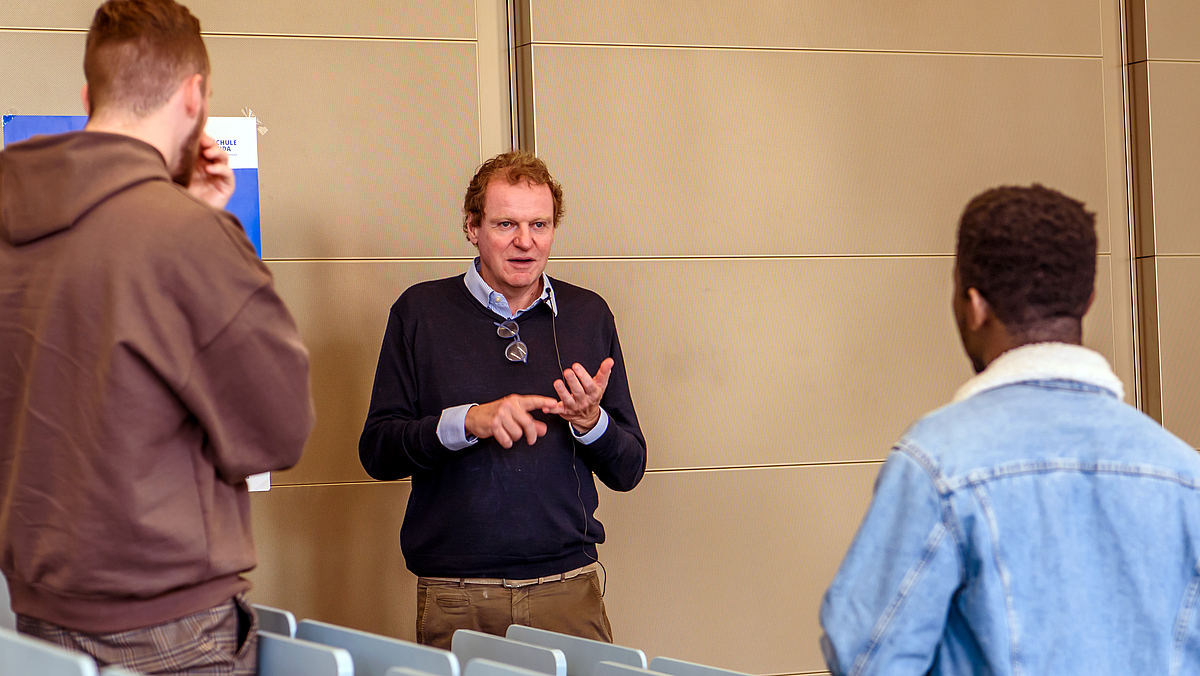 Dr. Franz Georg Strauß spricht mit zwei Studierenden, die mit viel Abstand vor ihm stehen. Er gestikuliert, tippt mit dem rechten Zeigefinger auf den kleinen Finger der linken Hand.