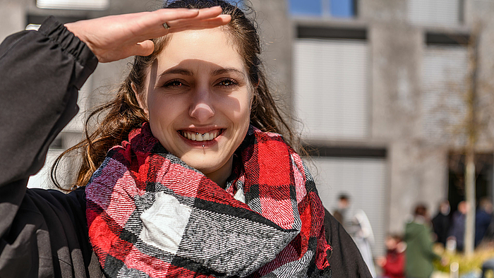 Das Foto zeigt eine lachende junge Frau. Sie hält ihre Hand an die Stirn, um nicht geblendet zu werden. Im Hintergrund ist unscharf das Zentrum für Medien und Soziale Arbeit der Hochschule Mittweida zu sehen. 