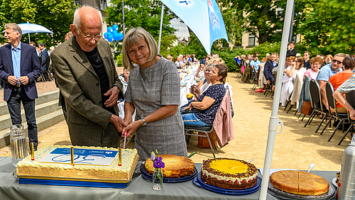 Das Foto zeigt eine männliche und eine weibliche Person. Sie fassen gemeinsam ein Messer an und schneiden eine Torte vor sich auf einem Tisch an, auf der eine große 30 zu erkennen ist. Im Hintergrund sind zahlreiche Menschen an langen Tafeln sitzend zu sehen. 