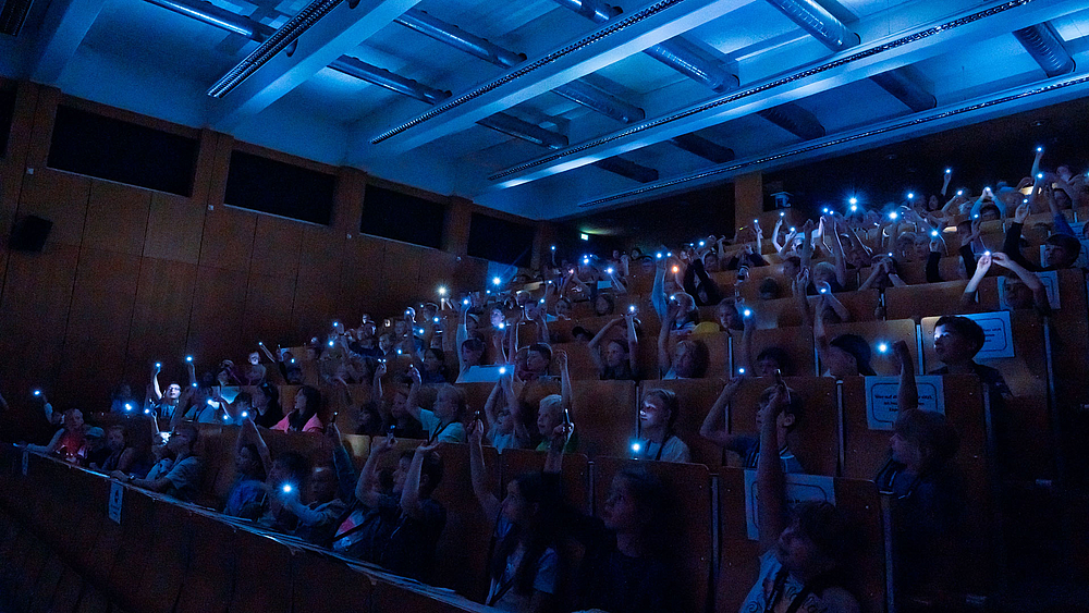 Großer Hörsaal voller Kinder, die im abgedunkelten Raum gemeinsam kleine Taschenlampen hochhalten.