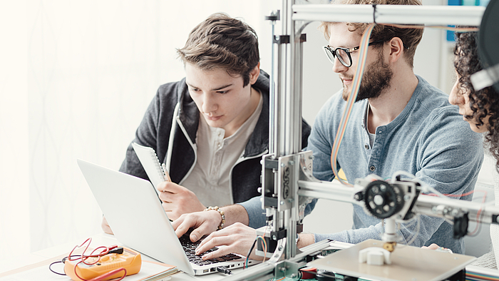 Zwei junge Männer sitzen vor einem Laptop. Neben ihnen steht ein 3D-Drucker, der ein Plastikteil herstellt.