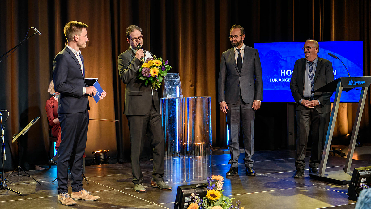 Auf dem Foto sind fünf Personen auf der Bühne unmittelbar nach der Übergabe zu sehen: Moderator Max Benda, Preisträger Marc Ritter, Staatsminister Sebastian Gemkow und Rektor Ludwig Hilmer.