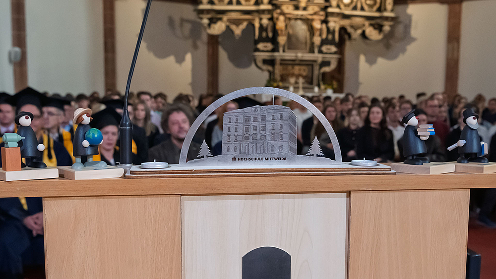 Blick über das Rednerpult mit Hochschuldevotionalen auf die dahinter sitzeenden Professor:innen und Erstsemester.
