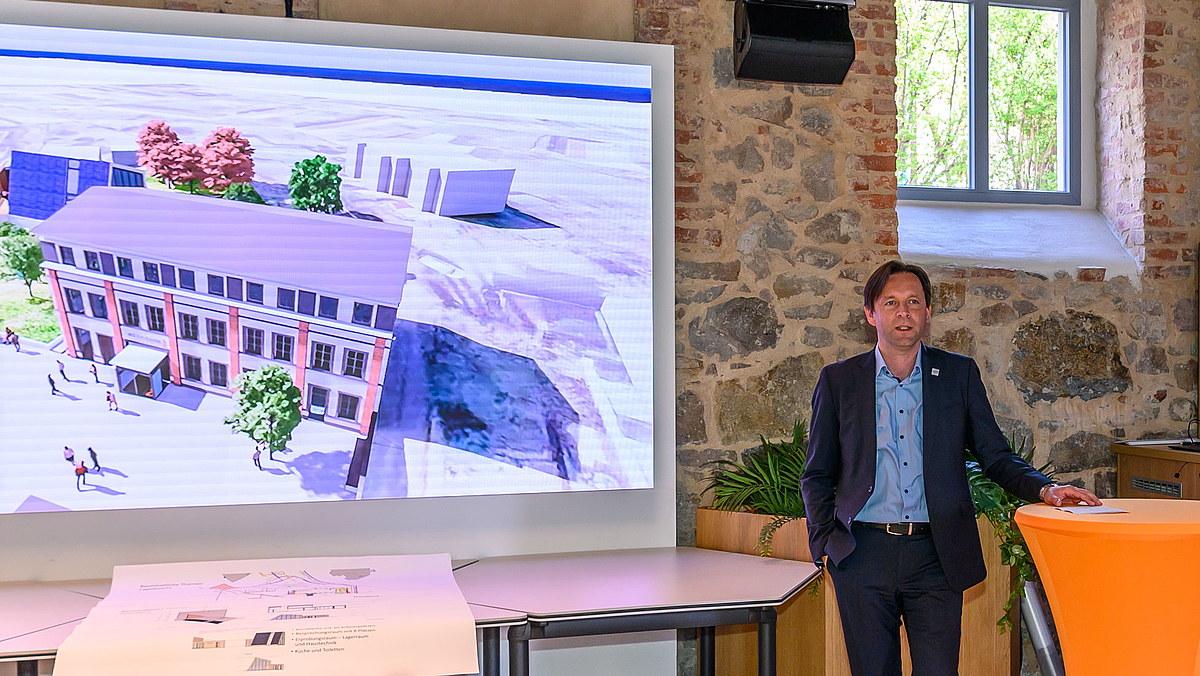 „Mittweida ist ein Paradebeispiel für innovationsgestützte Regionalentwicklung“, lobt Ronny Zienert vom SMR die Zusammenarbeit von Wissenscha Das Foto zeigt eine männliche Person an einem Stehtisch. Auf dem Bild links daneben ist auf einem großen Display die Animation des Werkbank32-TELEWERK-Geländes zu sehen.