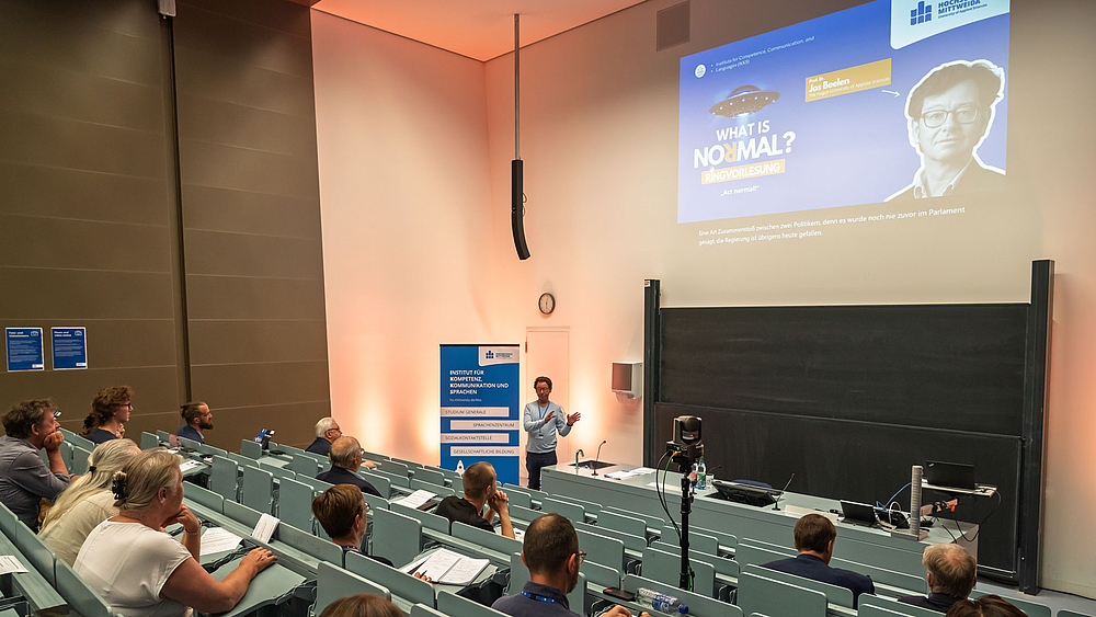 Blick über die Köpfe des Publums hinweg auf den männlichen Dozenten am unteren Ende des Hörsaals. HInter ihm steh ein blaues Rollup des IKKS und über ihm ist ine Folie projiziert mit dem Logo der Reihe "Was ist normal?" und Titel der Vorlesung "Act normal!".