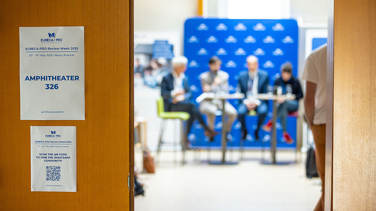 Durch eine offene Tür sieht man vier Personen, die auf einem Panel miteinander reden. Am linken Türflügel hängt ein Schild mit der Aufschrift "Autitorium".
