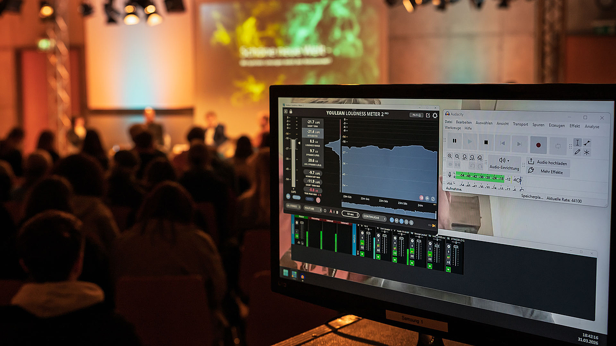 Im Vordergrund ein Monitor, der die Informationen eines digitalen Tonmischpults zeigt. Im Hintergrund ist unscharf der Saal mit Zuschauenden und dem Podium zu erkennen.