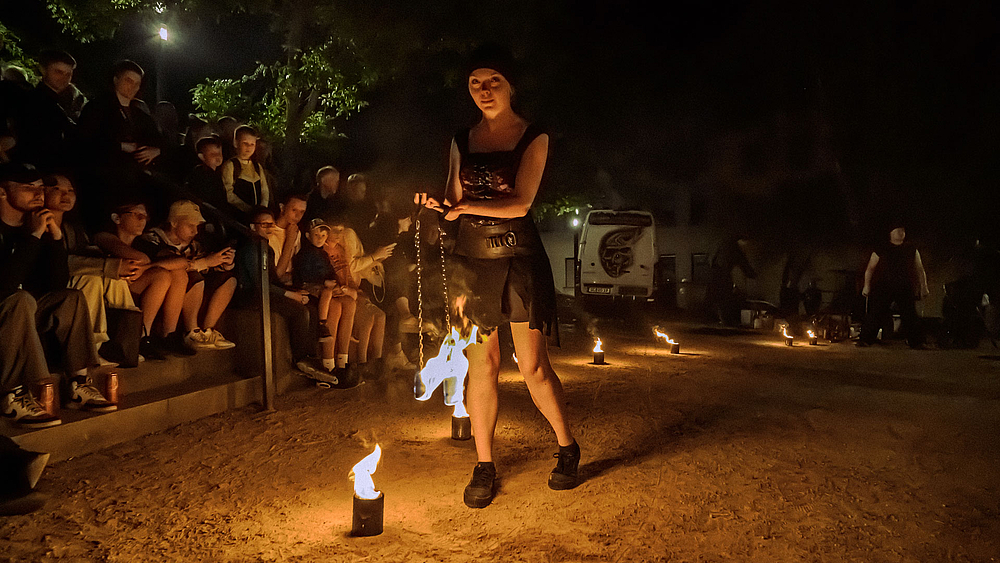 Eine junge weibliche Preson in schwarzre Kleidung hantiert auf einem Platz mit zwei Feuer-Pois. Auf dem Boden sind kleine Feuereschalen verteilt. Am linken Bildrand sind Zuschauer zu sehen.