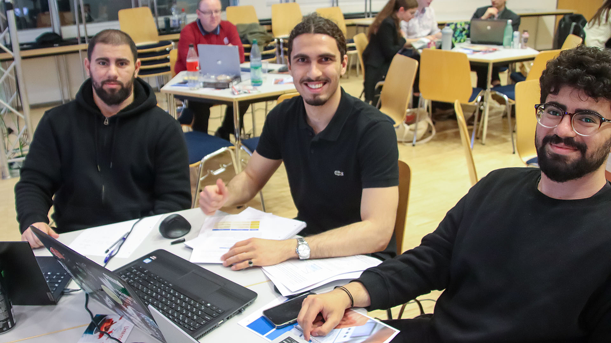 Drei junge männliche Personen mit Bärten an einem Tisch mit Laptops und zahlreichen Papieren schauen lächelnd in Richtung Kamera.