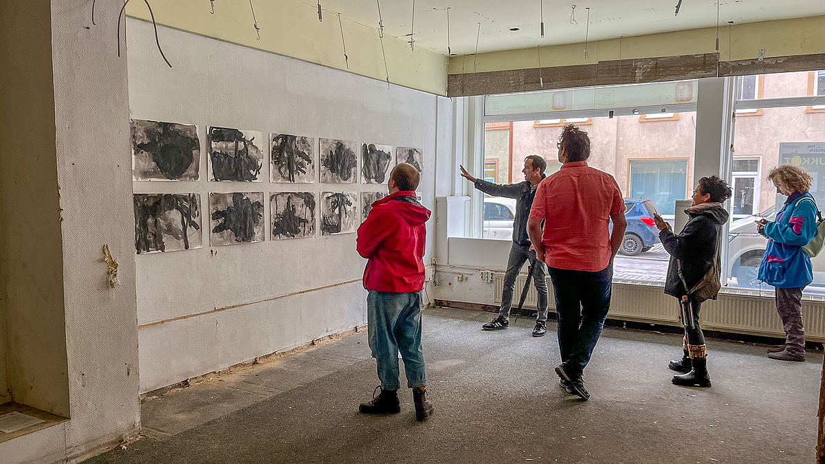 Ein traumatisch wirkende Installation aus Papierbögen an der Wand eines leerstehenden Ladens. Personen im Laden betrachten die Blätter.