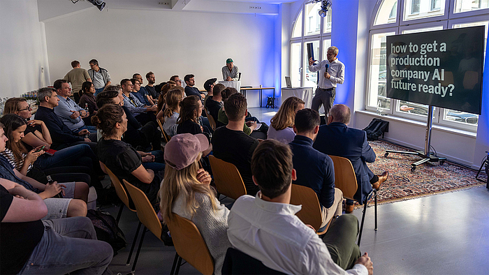 Ein Mann mit grauen Haaren und blauem Business-Hemd spricht gestikulierend vor einer im Halbkreis in mehreren Reihen sitzenden Gruppe.