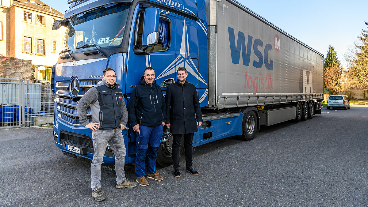 Das Bild zeigt drei Männer an der Mercedes-Zugmaschine eines großen Sattelzuges stehen. Der dreiachsige Auflieger trägt auf der Plane den Schriftzug „WSG Logistik