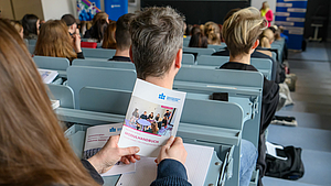 Blick von hinten auf eine A5-Broschüre mit dem Titel „Modulhandbuch“ in den Händen einer weiblichen Person, die in eine Hörsaalbankreihe sitzt.