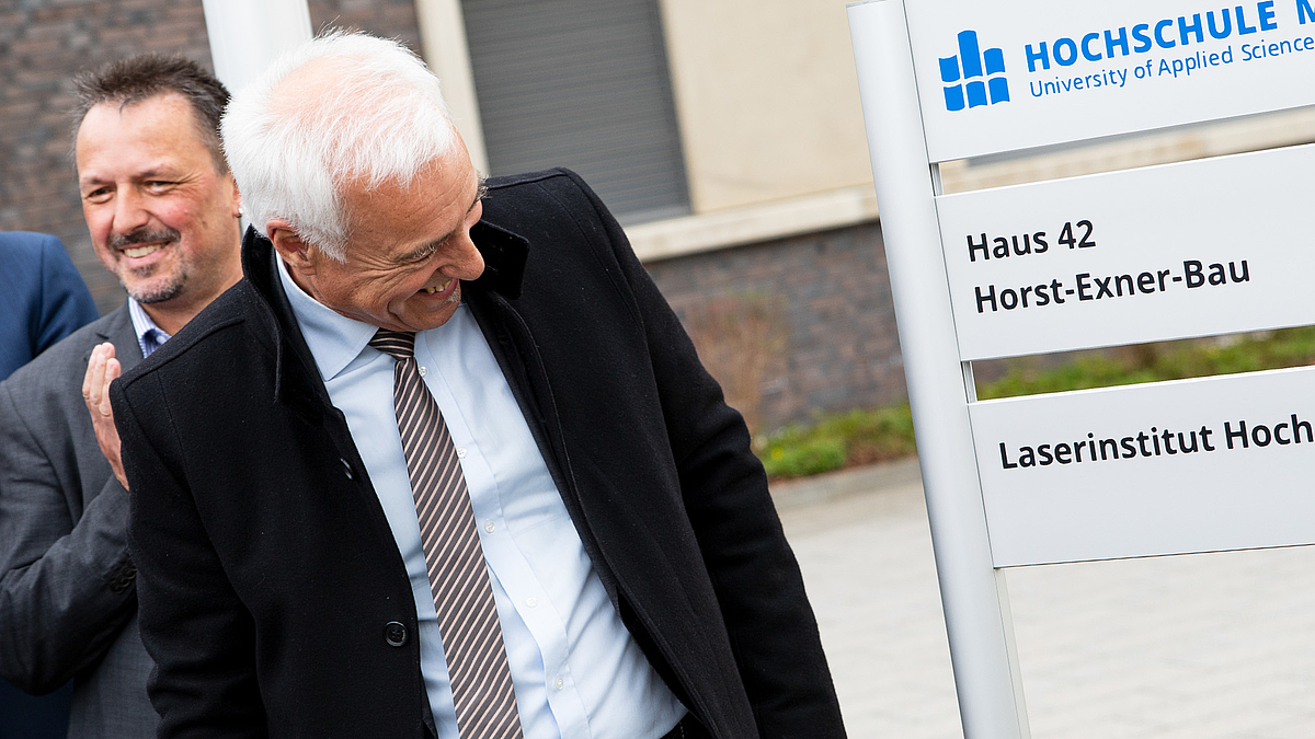 Professor Horst Exner blickt lächelnd auf das Schild mit der Aufschrift "Horst-Exner-Bau" vor dem Laserinstitut Hochschule Mittweida. Im Hintergrund applaudiert Professor Udo Löschner.