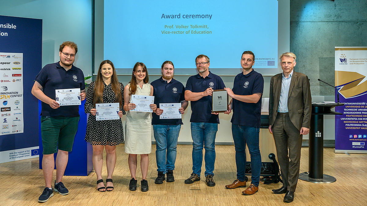 Sechs Personen des Teams mit dem 1. Platz stehen nebeneinander und halten Urkunden bzw. Preissymbole in den Händen. Rechts von ihnen seht eine weitere männliche Person. Alle blicken in die Kamera.