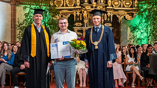 Drie maännliche Personen stehend im Altarrarum einer Kirche. Die jünger Person in der Mitte hält einen Blumenstrauß und eine Urkunde in den Händen. Die Personen links und rechts tragen akademsiche Talare und Hüte, die Person rechts zusätzllich eine goldene Amtskette.