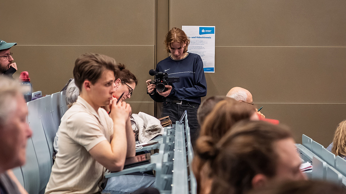Blick durch die besetzten ansteigenden Banhkreihen im Hörsaal auf eine weibliche Person mit einer professionellen Videokamera in der Hand.