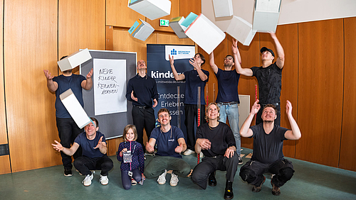 Auch das Team hinter der Kinderuni sagt: Tschüss Schubladendenken! Schubladen helfen zwar beim Ordnen des Alltags, aber in unseren Köpfen sorgen sie oft für falsche Bilder – und Vorurteile. Eine Gruppe von zehn Personen im Hörsaal, die Schubladen aus Karton in die Luft werfen und wieder auffangen. Im Hintergrund sieht man ein dunkelblaues Kinderuni-Banner und eine Stellwand mit der Aufschrift „Neue Kinder kennenlernen“.