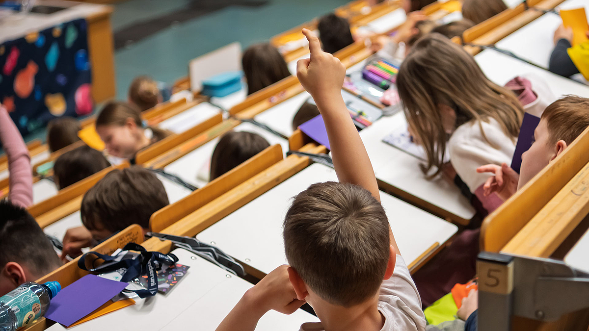 Kind im Hörsaal streckt seinen reichten Arm nach oben und meldet sich.