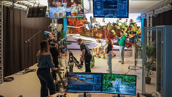 Junge Menschen im Studioraum mit Kameras, Scheinwerfen udn Monitoren. Im Bildhintergrund sitzen zwei Oersonen in Liegestühlen vor einer raumgroßen LED-Wand, die einen Palmenstrand zeigt.