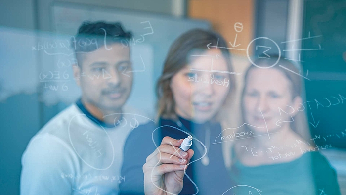 Blick durch eine Glasscheibe auf drei junge Menschen, ein Mann links, daneben zwei Frauen. Die junge Frau in der Mitte schreibt mit einen weißen Stift auf die Glasscheibe mathematische Formeln..